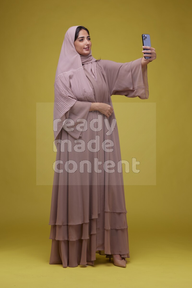 A woman taking a Selfie on a Yellow Background wearing Brown Abaya with Hijab