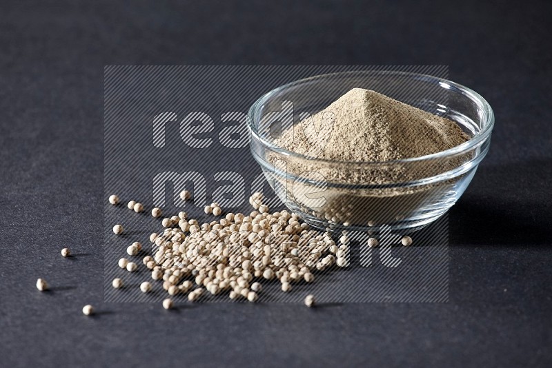 A glass bowl full of white pepper powder with white pepper beads on black flooring