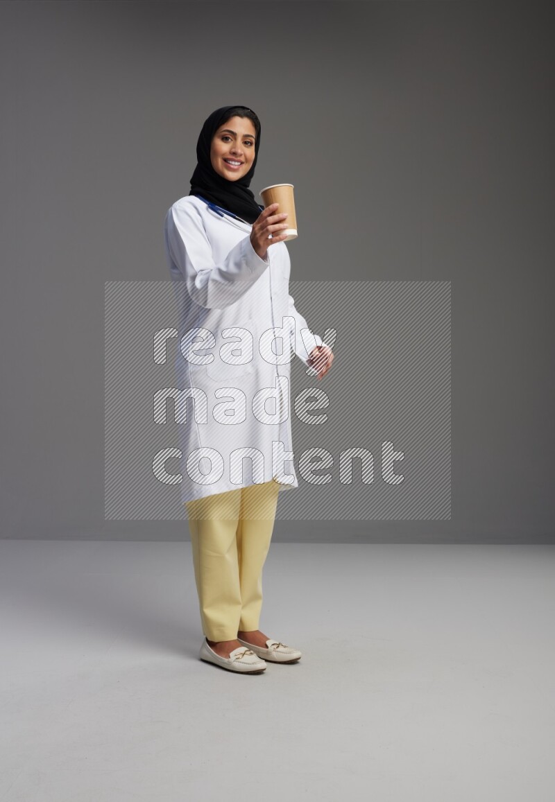 Saudi woman wearing lab coat with stethoscope standing holding paper cup on Gray background