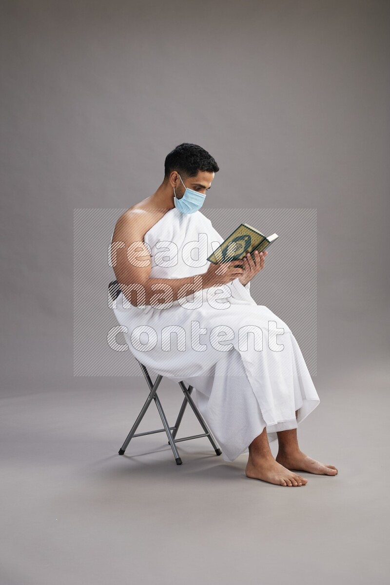 A man wearing Ehram with face mask sitting on chair reading quran on gray background