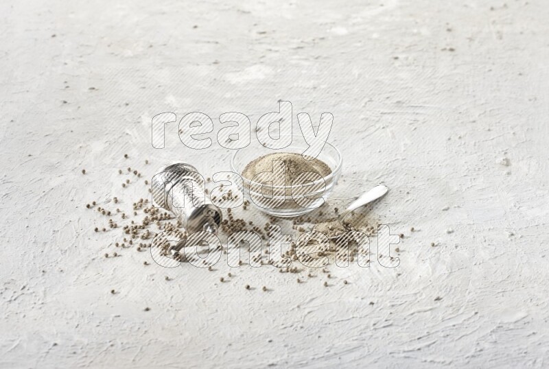 A glass bowl full of white pepper powder with white pepper beads and a metal grinder on textured white flooring