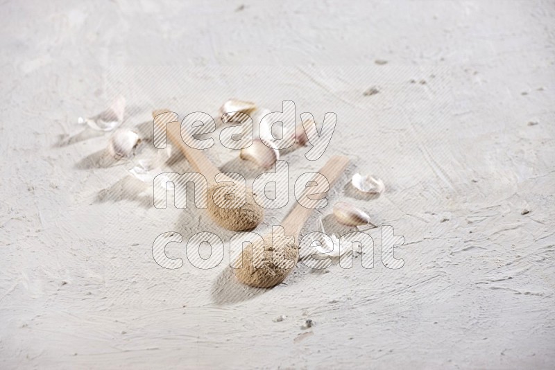2 wooden spoons full of garlic powder with cloves and peels on a textured white flooring in different angles
