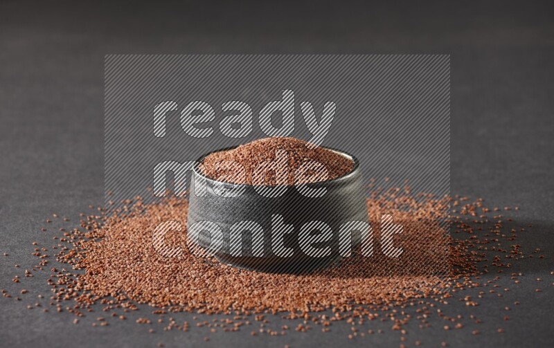 A black pottery bowl full of garden cress seeds surrounded by seeds on a black flooring