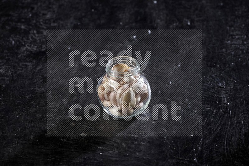 A glass spice jar full of garlic cloves on a textured black flooring in different angles