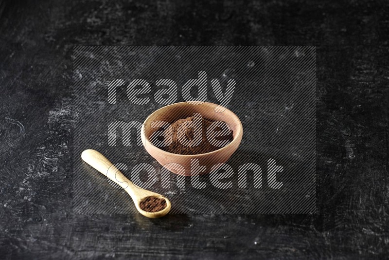 A wooden bowl and wooden spoon full of cloves powder on textured black flooring