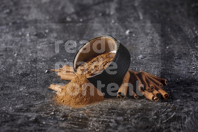 Black pottery bowl over filled with cinnamon powder and cinnamon sticks around the bowl on a textured black background