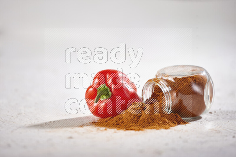 A glass jar full of ground paprika powder flipped with some spilling powder on white background