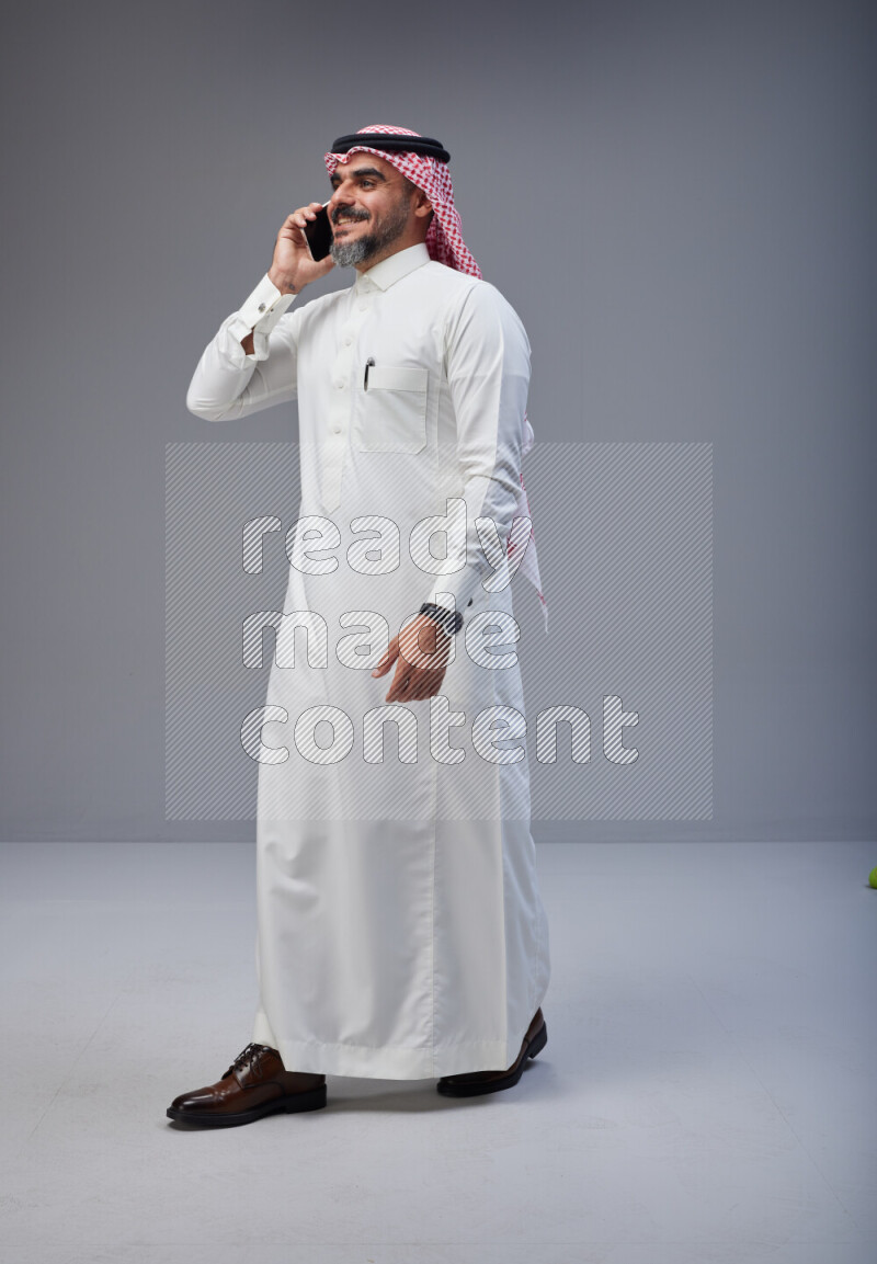 Saudi man Wearing Thob and red Shomag standing talking on phone on Gray background