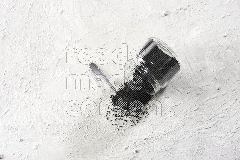 A glass jar and a metal spoon full of black seeds and jar is flipped and seeds spread on a textured white flooring in different angles