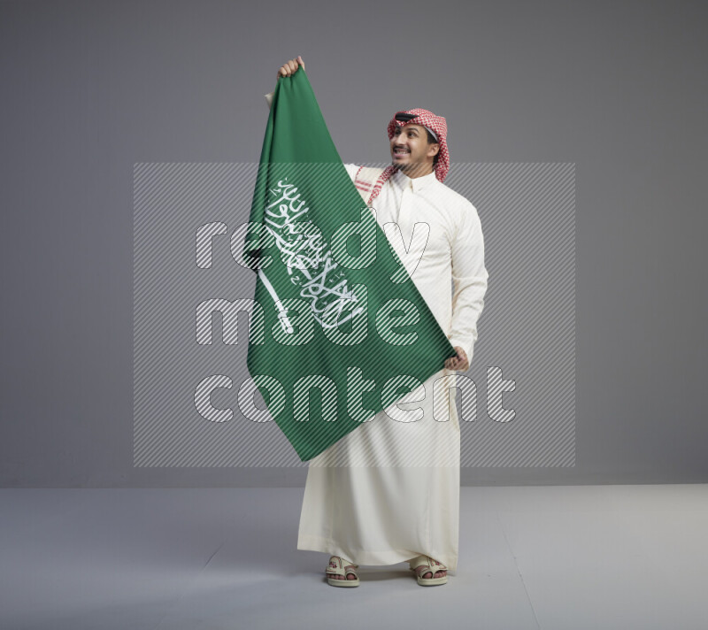 A saudi man standing wearing thob and red shomag holding big saudi flag on gray background