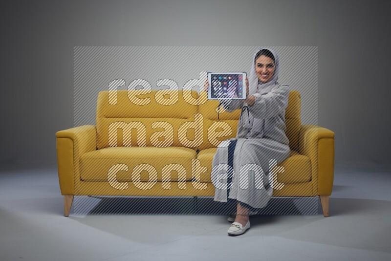 A Saudi female wearing a light gray Abaya and head scarf sitting on a yellow sofa and showing her tablet's screen while wearing headphones eye level on a grey background