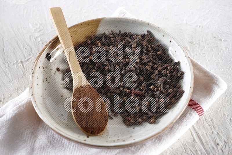 A Pottery plate full of whole cloves and a wooden spoon full of cloves powder in it on a textured white background