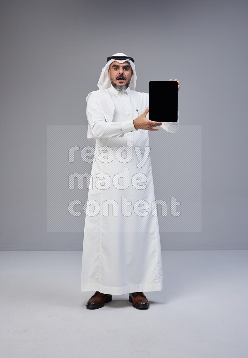 Saudi man Wearing Thob and white Shomag standing showing tablet to camera on Gray background