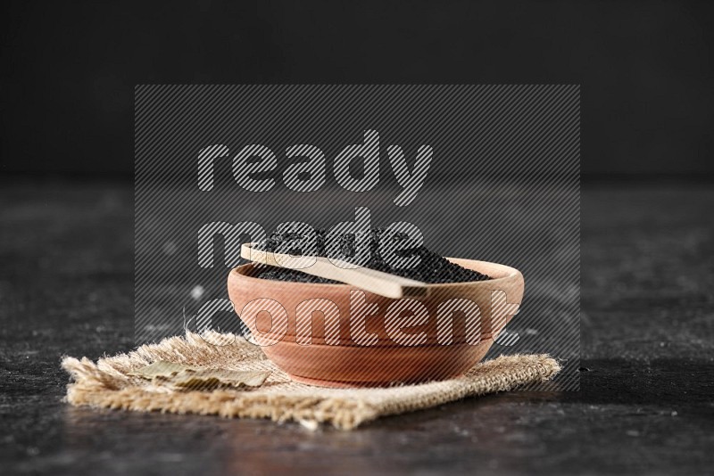 A wooden bowl full of black seeds with wooden spoon full of the seeds on it on a burlap fabric on a textured black flooring