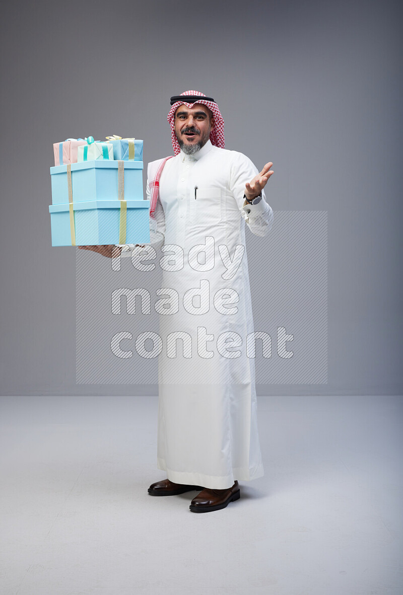Saudi man Wearing Thob and red Shomag standing holding gift box on Gray background