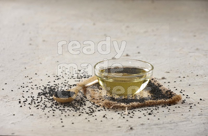 A glass bowl full of black seeds oil and wooden spoon full of black seeds with seeds spread on burlap fabric on a textured white flooring