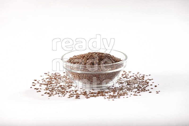 A glass bowl full of flax seeds surrounded by flax seeds on a white flooring