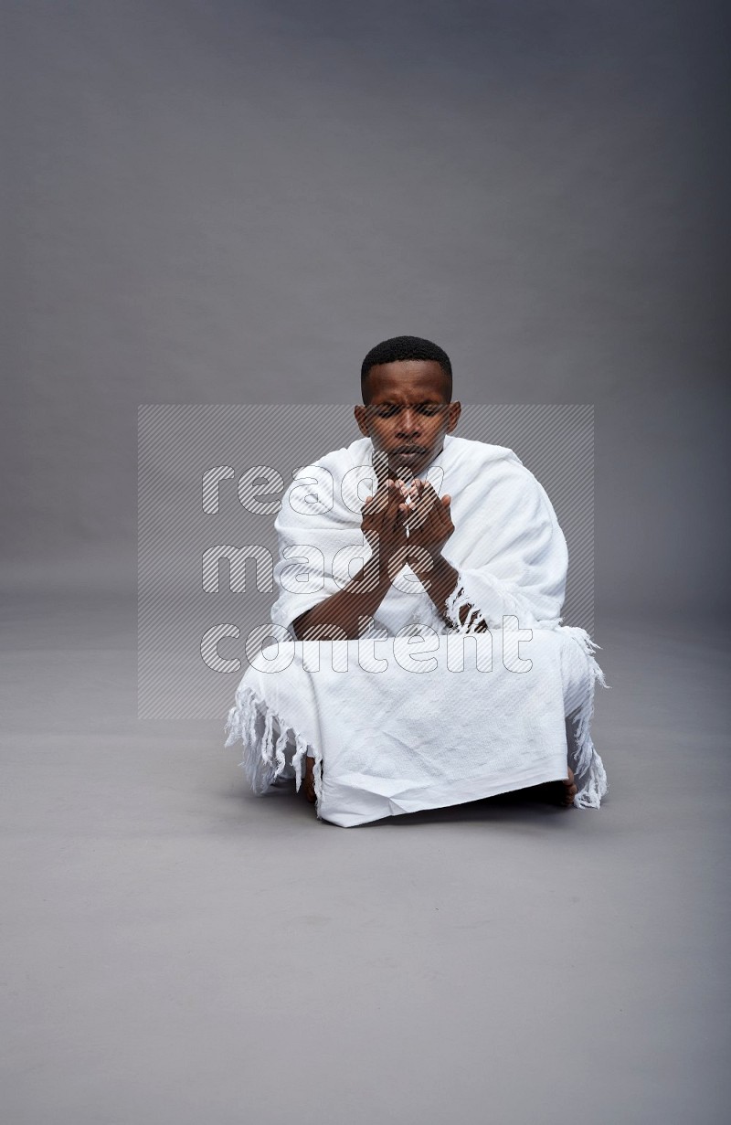 A man wearing Ehram sitting on floor dua'a on gray background