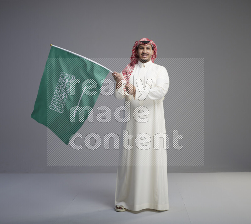 A Saudi man standing wearing thob and red shomag raising big Saudi flag on gray background