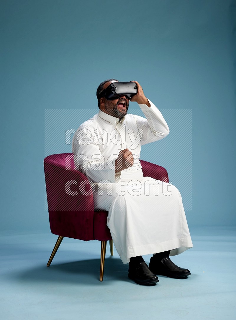 Saudi Man without shimag sitting on chair  gaming with VR on blue background