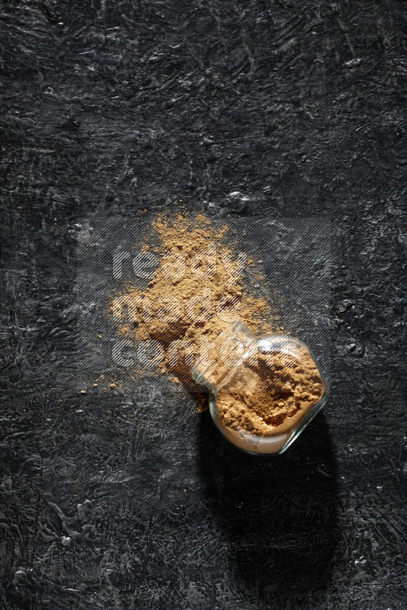 A flipped glass spice jar full of allspice powder and powder spilled out of it on a textured black flooring