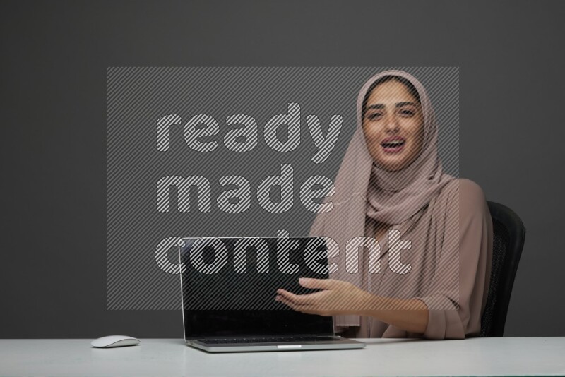 A woman Sitting on her desk  Pointing at her laptop on a Gray Background wearing Brown Abaya with Hijab