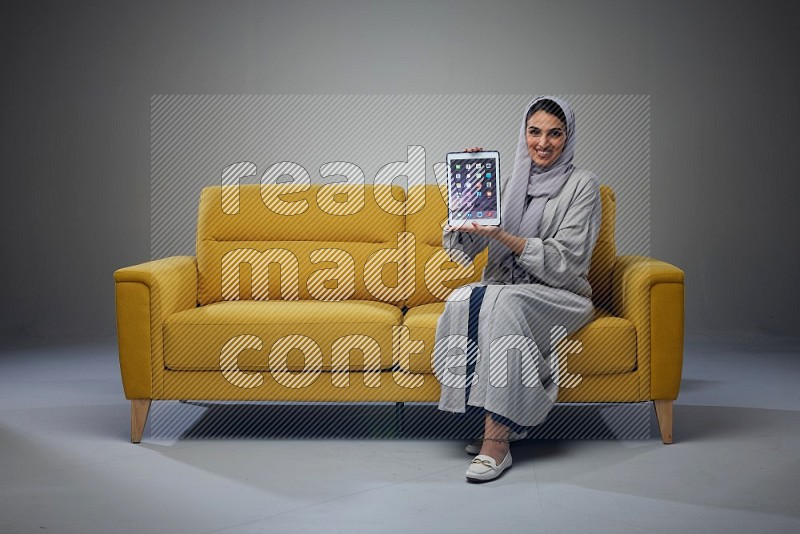 A Saudi female wearing a light gray Abaya and head scarf sitting on a yellow sofa and showing her tablet's screen while wearing headphones eye level on a grey background