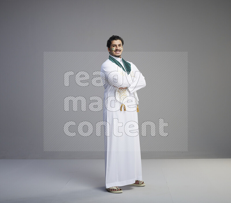 A Saudi man standing wearing thob and Saudi flag scarf with face painting on gray background