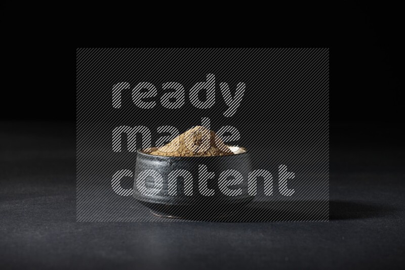 A black pottery bowl full of cumin powder on a black flooring