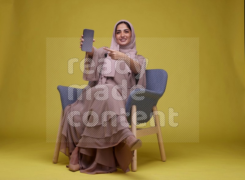 A woman Showing Her Phone Screen Setting  on a Yellow Background wearing Brown Abaya with Hijab