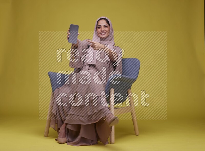 A woman Showing Her Phone Screen Setting  on a Yellow Background wearing Brown Abaya with Hijab