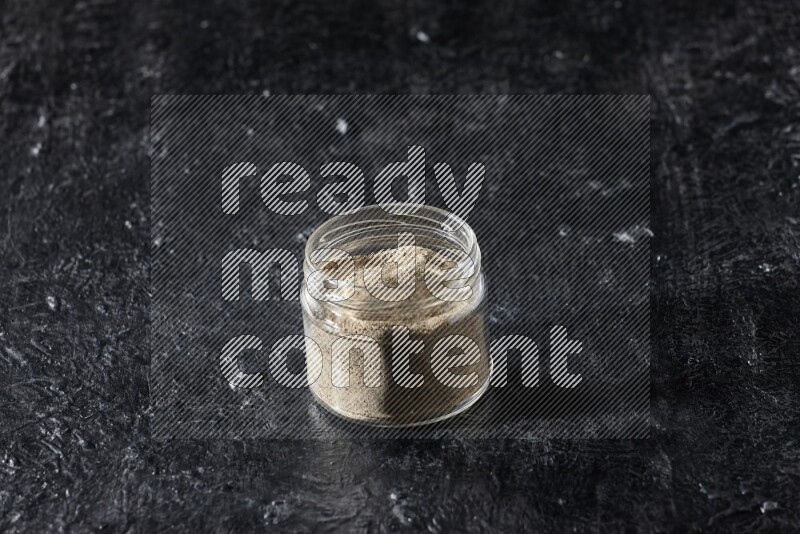A glass jar full of white pepper powder on textured black flooring