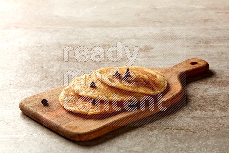 Three stacked chocolate chips pancakes on a wooden board on beige background