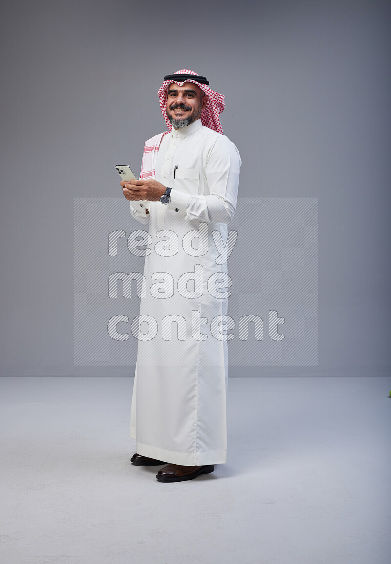 Saudi man Wearing Thob and red Shomag standing texting on phone on Gray background