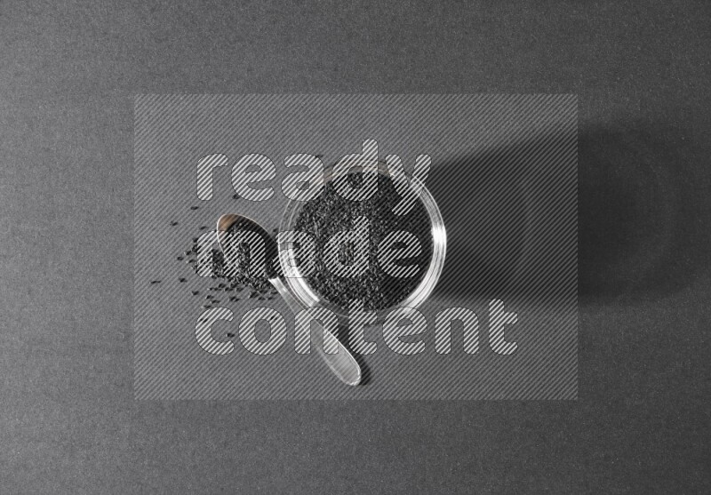 وعاء زجاجي ممتلئ ببذور حبة البركة مع ملعقة معدنية مليئة بحبة البركة علي خلفية سوداء