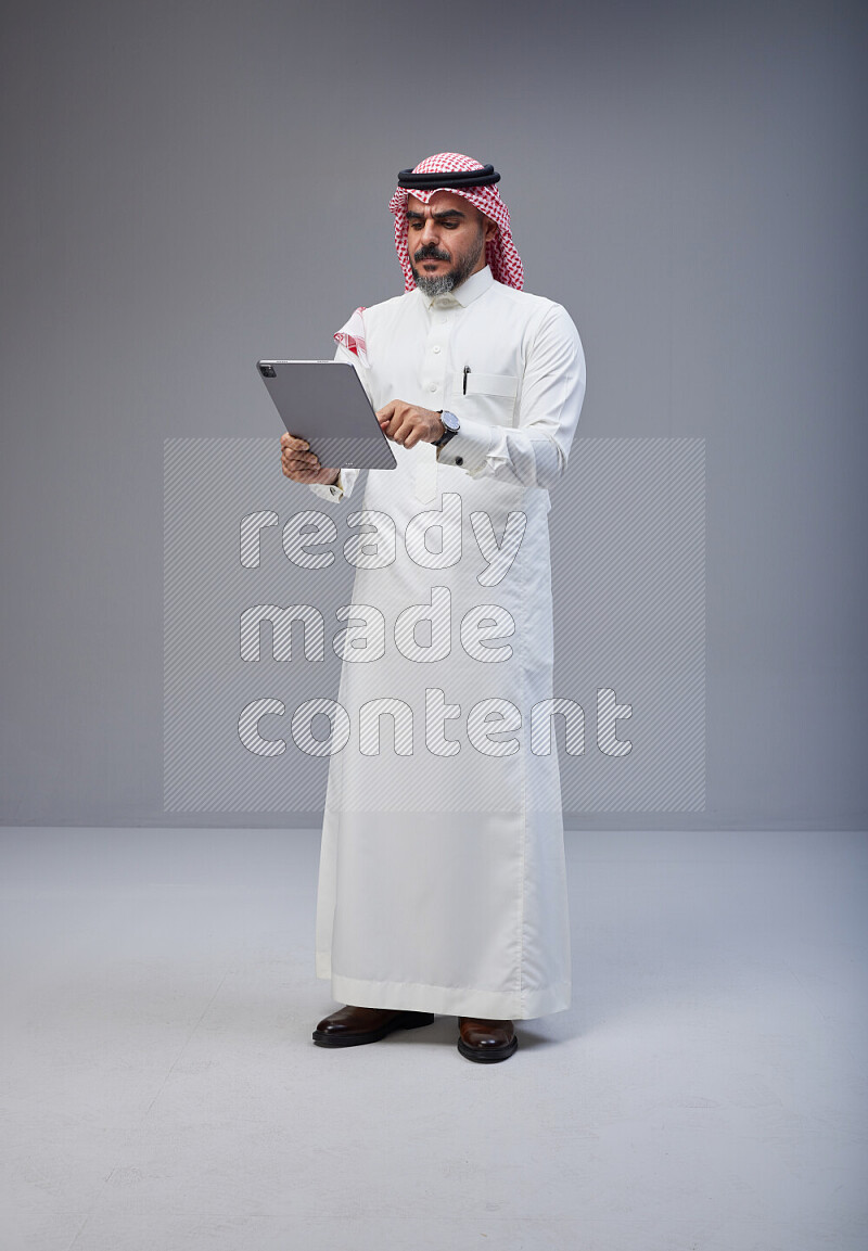 Saudi man Wearing Thob and red Shomag standing working on tablet on Gray background
