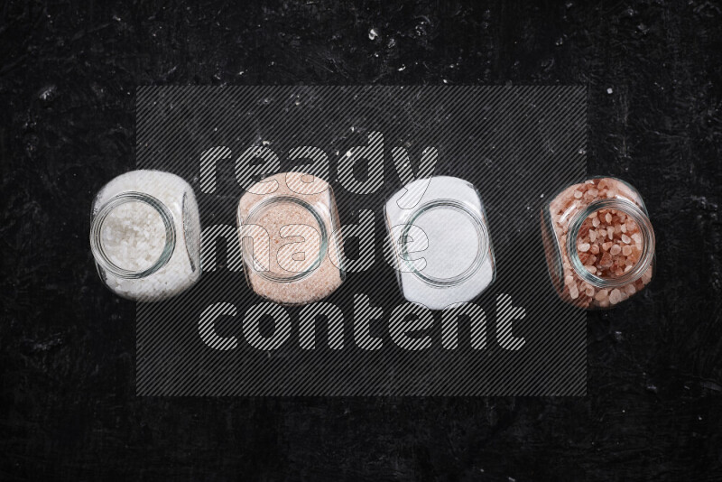 4 glass jars filled with table salt, coarse sea salt, fine himalayan salt and coarse himalayan salt on black background