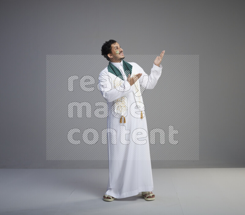 A Saudi man standing wearing thob and Saudi flag scarf with face painting on gray background