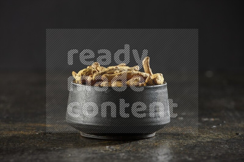 A black pottery bowl full of dried turmeric whole fingers on textured black flooring