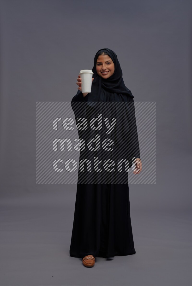 Saudi woman wearing Abaya standing holding paper cup on gray background