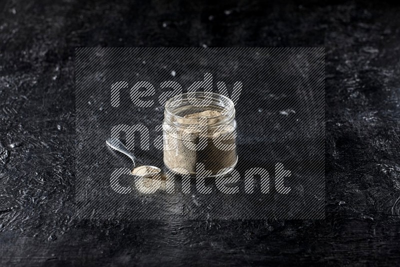 A glass jar and a metal spoon full of white pepper powder on textured black flooring