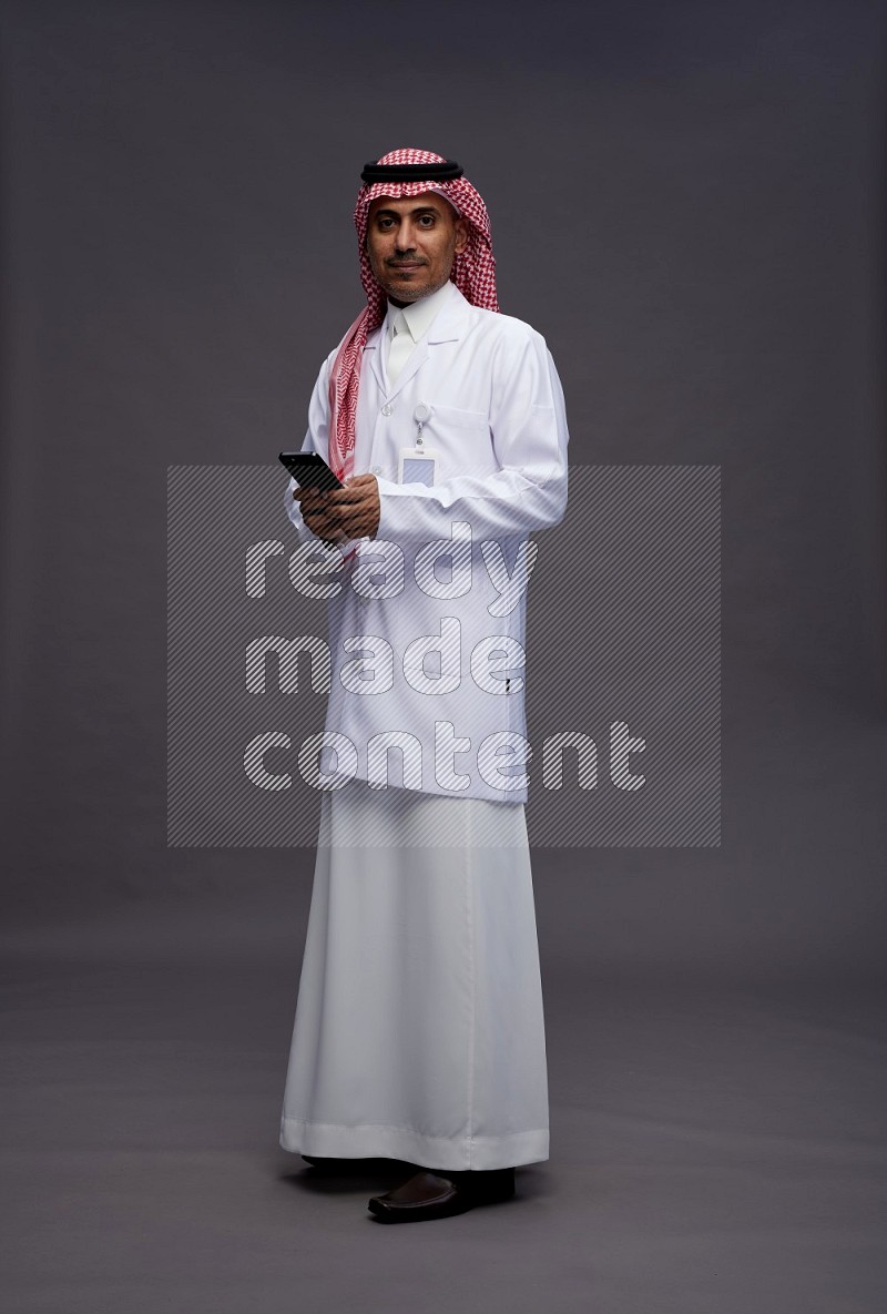 Saudi man wearing thob with lab coat and shomag with pocket employee badge standing texting on phone on gray background