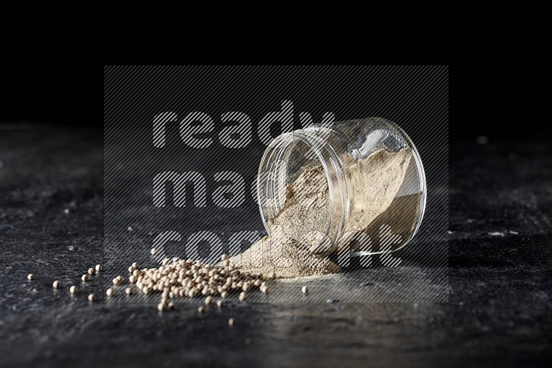 A flipped glass jar full of white pepper powder with spilled powder and pepper beads on textured black flooring