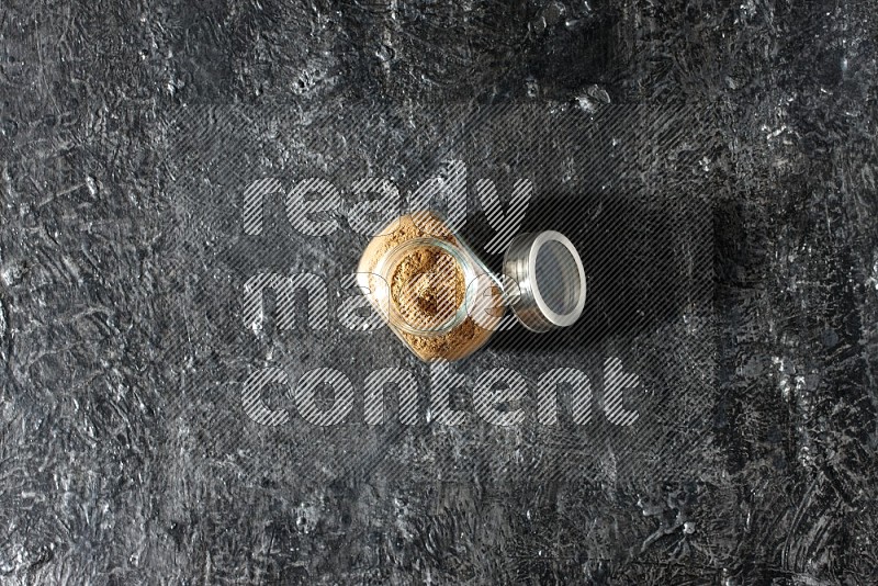 A glass spice jar full of allspice powder on a textured black flooring