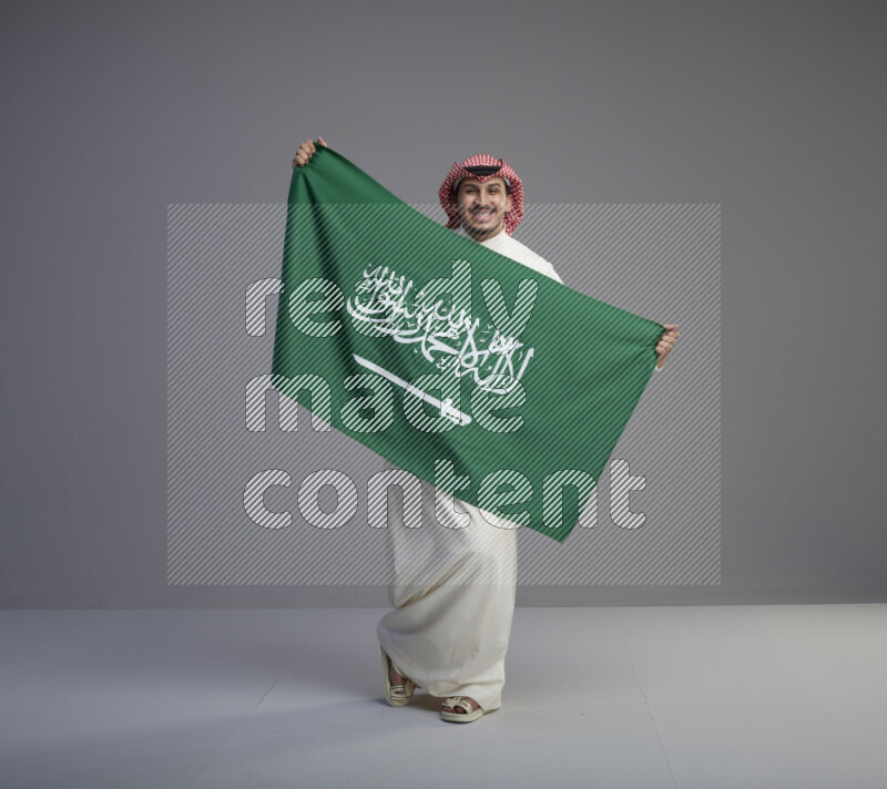 A saudi man standing wearing thob and red shomag holding big saudi flag on gray background