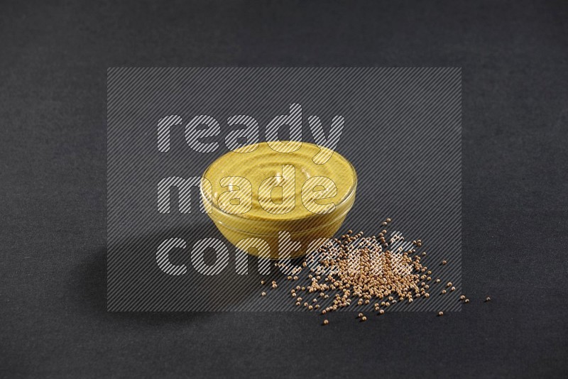 A glass bowl full of mustard paste with mustard seeds underneath on black flooring