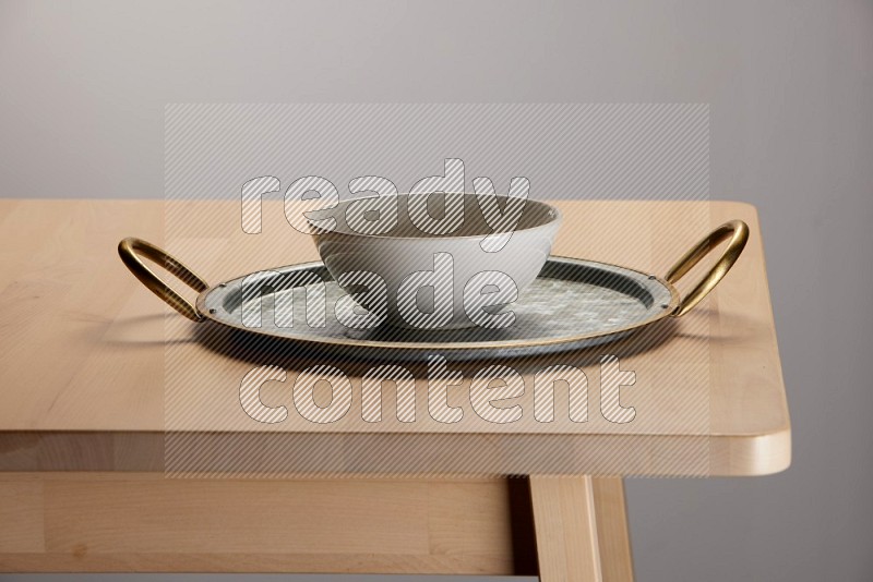 off white bowl with islamic pattern placed on a rounded stainless steel tray with golden handels on the edge of wooden table