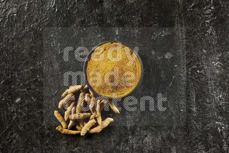 A black pottery bowl full of turmeric powder and dried turmeric whole fingers next of it on textured black flooring