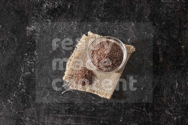 A glass bowl full of flaxseeds with bunch of the seeds on burlap fabric on a textured black flooring