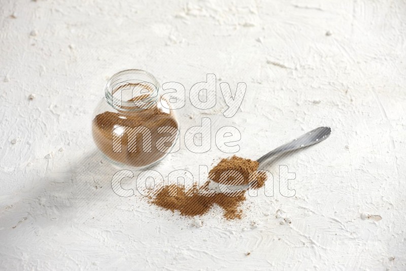 Herbs glass jar full of cinnamon powder with a metal spoon full of powder on a textured white background